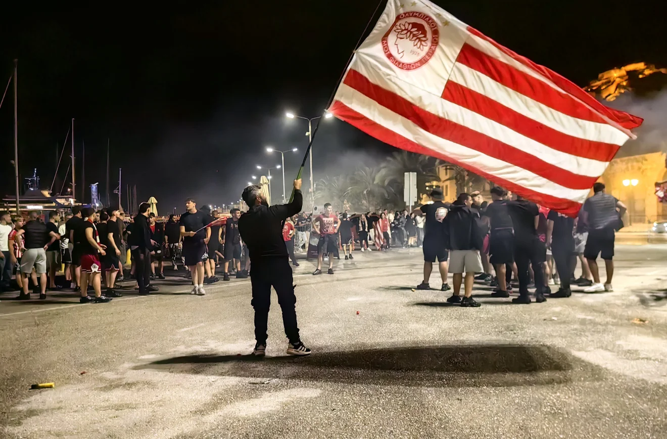 Οπαδός του Ολυμπιακού ανεμίζει μεγάλη ερυθρόλευκη σημαία σε νυχτερινή συγκέντρωση φιλάθλων, με καπνούς και κόσμο στο φόντο. Πηγή: ΙΝΤΙΜΕ.