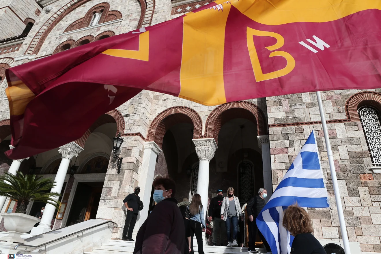 Πιστοί έξω από ιερό ναό, με εκκλησιαστικές σημαίες και την ελληνική σημαία να κυματίζουν μπροστά από την είσοδο. Πηγή: ΙΝΤΙΜΕ.