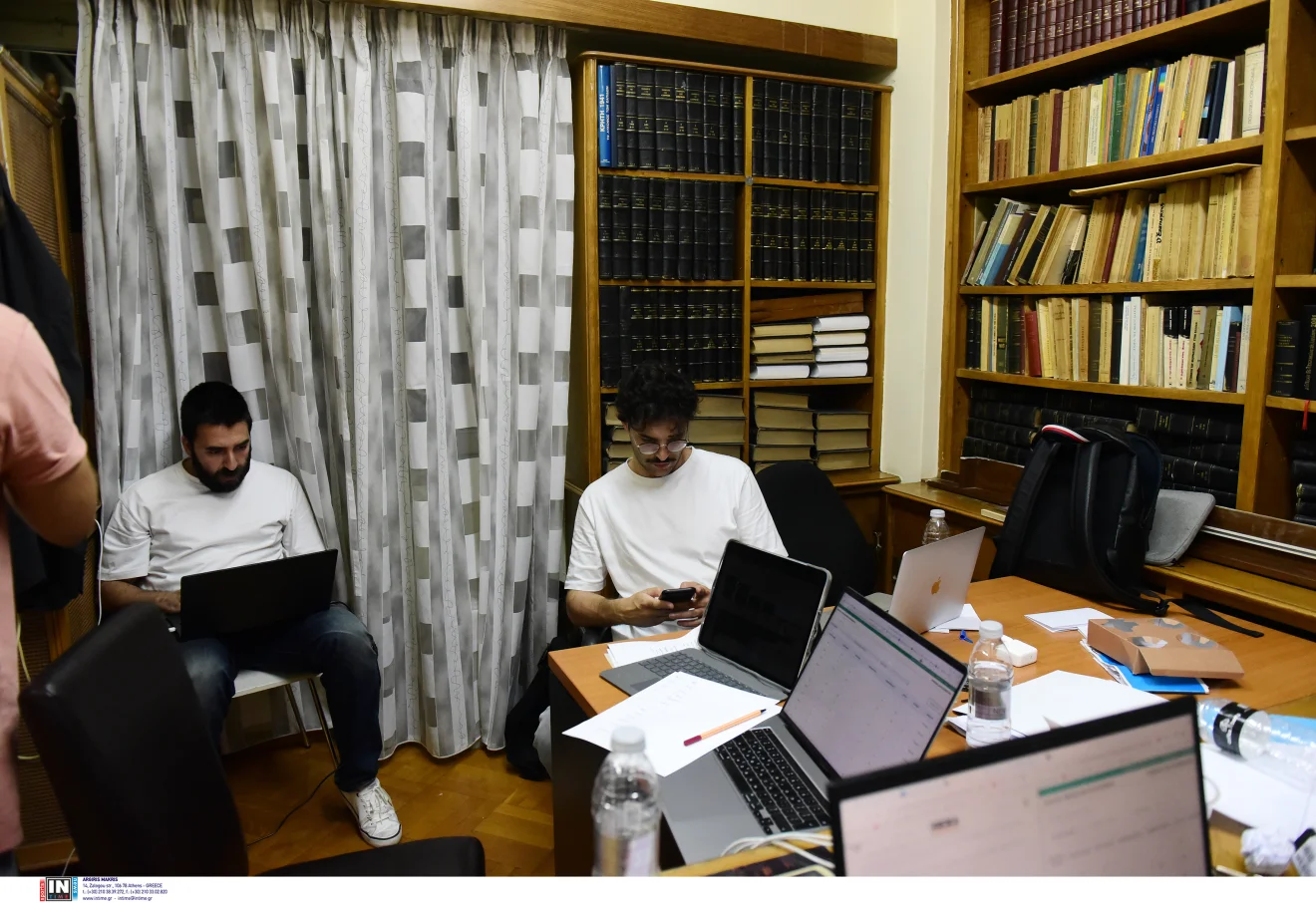 Δύο άνδρες εργάζονται με φορητούς υπολογιστές και κινητά τηλέφωνα σε γραφείο με μεγάλες βιβλιοθήκες και πολλά βιβλία. Πηγή: INTIME.
