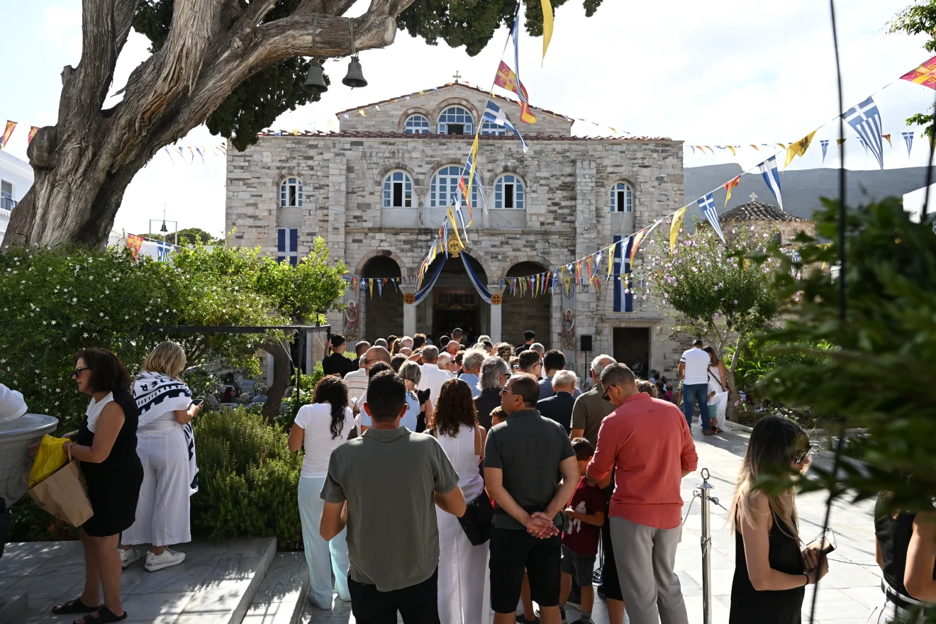 Πλήθος πιστών έξω από Ιερό Ναό, με ελληνικές και βυζαντινές σημαίες να πλαισιώνουν τον χώρο κατά τη διάρκεια θρησκευτικού εορτασμού. Πηγή: ΙΝΤΙΜΕ